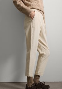 Suéter beige texturizado con diseño de punto de cable, combinado con pantalones cortos color crema que cuentan con bolsillos laterales y puños doblados. Zapatos y calcetines marrones.