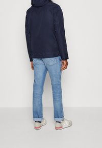 Navy jacket with a hood, smooth texture. Light blue jeans, rolled cuffs. Neutral-colored sneakers with three stripes and a red accent.