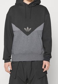 Svart och grå färgblockerad huvtröja med Adidas trefoil-logo på bröstet, bärs av en person med avslappnade händer vid sidorna.
