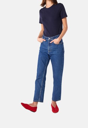 Femme portant un t-shirt à manches courtes bleu marine, un jean taille haute bleu et des chaussures plates rouges, debout les mains dans les poches sur un fond blanc.