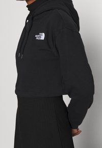 Krátká černá mikina s kapucí z bavlněné směsi, vybavená přední kapsou, stahovací šňůrkou na kapuci a bílým logem na hrudi. Jemná textura.