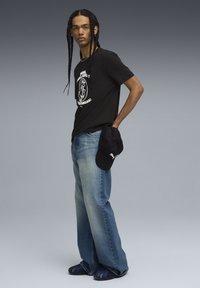 Black graphic t-shirt, light blue wide-leg jeans, and black cap. Braided hair and sandals with blue accents. Neutral gradient background.