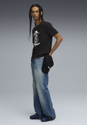 Sort grafisk t-shirt, lyseblå vide bukser og sort kasket. Flettet hår og sandaler med blå detaljer. Neutral gradient baggrund.