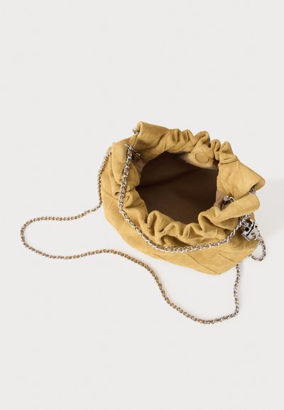 Petit sac en suède beige doux avec cordon coulissant et bandoulière chaîne argentée, ouverture supérieure montrant un intérieur marron vide sur fond clair uni.