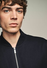 Chaqueta bomber de color navy con cuello acanalado y detalle de cremallera frontal. El material parece texturizado y duradero, enfatizando un diseño moderno.