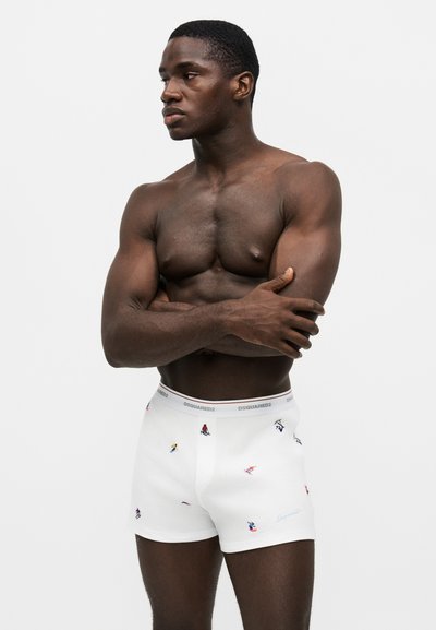 Homme musclé, bras croisés, portant un boxer blanc décoré de petits motifs colorés de skieurs sur un fond uni.