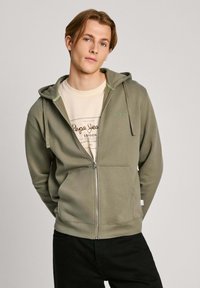 Sudadera con cremallera en verde oliva, con bolsillos frontales, acentos en verde claro y una textura suave, superpuesta sobre una camiseta beige con un estampado de logo.