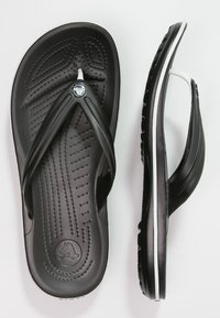 Svarta flip-flops med en texturerad innersula, tunna remmar och en vit accentlinje på sulan. Har en logotyp på remmen.