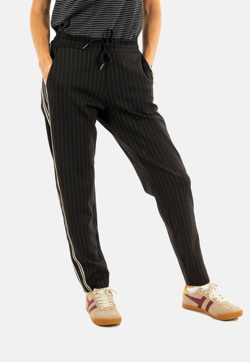 Zwarte joggerbroek met krijtstrepen, elastiche tailleband, zijzakken en witte zijstrepen; gedragen met tan sneakers met bordeauxrode accenten.