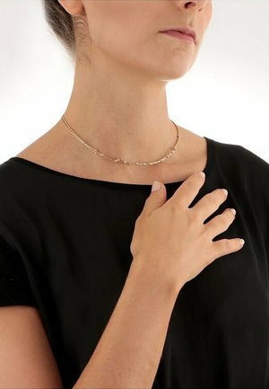 Goldfarbene Choker-Halskette mit kleinen Perlen, die ein zartes, geschwungenes Design aufweist. Am Hals getragen, passend zu einem schwarzen Oberteil.