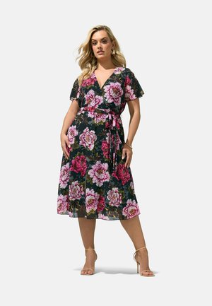 Frau trägt ein schwarzes Midi-Wickelkleid mit pinken und roten Blumenmustern, steht mit einer Hand in der Hüfte und trägt hautfarbene High-Heel-Sandalen.