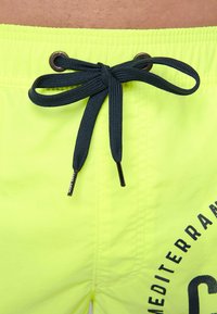 Pantaloni da bagno giallo neon in tessuto texturizzato, con coulisse blu scuro e dettagli grafici stampati sul lato.