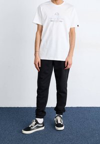 Valkoinen puuvillapaita, jossa on sininen ja harmaa graafinen teksti, yhdistettynä mustiin collegehousuihin ja mustiin lenkkareihin, joissa on valkoiset yksityiskohdat ja nauhat.