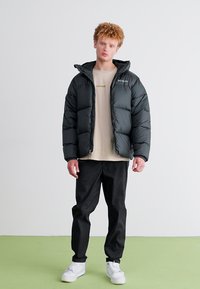 Schwarze, gepolsterte Jacke mit strukturierter Oberfläche, mit Kapuze und langen Ärmeln, getragen über einem beigen T-Shirt und schwarzen Hosen, ergänzt durch weiße Sneakers.