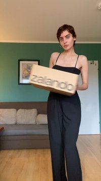 Kartonowe pudełko z napisem "zalando" drukowanym na szaro, trzymane przez osobę ubraną w czarny bezrękawnik, stojącą w pomieszczeniu z zielonymi ścianami.