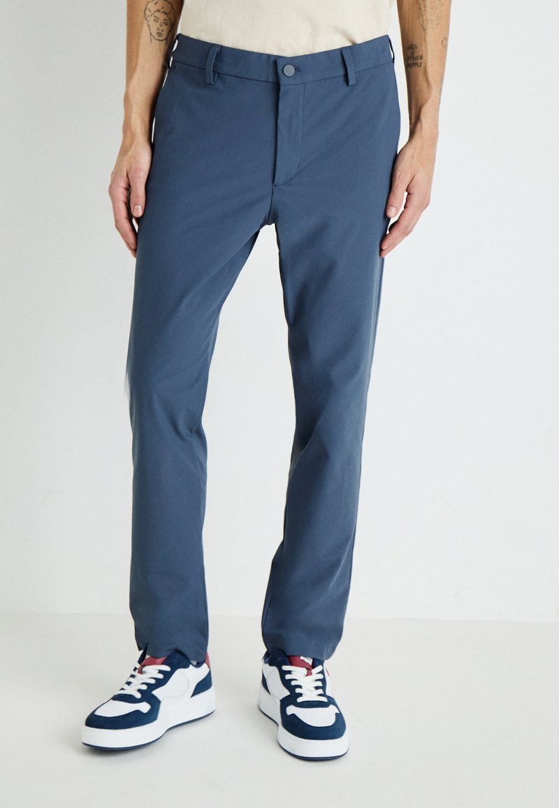 Men's navy blue pants made of smooth fabric, featuring a classic fit, side pockets, and a simple button closure. Paired with white and navy sneakers.