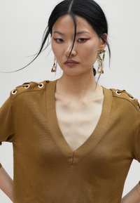 Top verde oliva a maniche corte con scollatura a V e dettagli sulle spalle. Il tessuto è liscio e aderente, con accenti dorati sulle spalle.