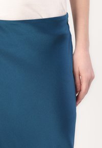 Gonna turchese realizzata in tessuto liscio, con un dettaglio di bottoni laterali. Design aderente con una leggera texture e una silhouette dritta.