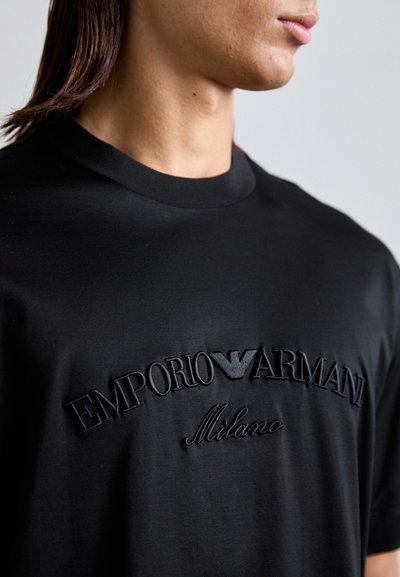 Persoană îmbrăcată cu un tricou negru cu text brodat în relief "EMPORIO ARMANI Milano" și logo-ul unui vultur pe piept.