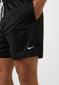 Sorte sports korte bukser i mesh med hvid snor og hvid Nike swoosh-logo, båret af en person med hånden i lommen.