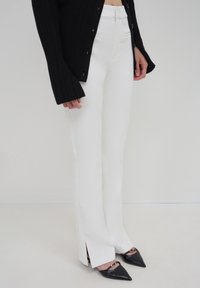 Donna che indossa pantaloni bianchi a vita alta con spacchi laterali, scarpe basse nere a punta con dettagli dorati e una blusa nera a maniche lunghe con bottoni.