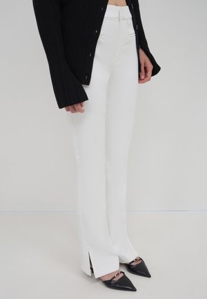 Woman wearing high-waist white pants with side slits, black pointed-toe flats with gold detail, and a black long-sleeve buttoned top.