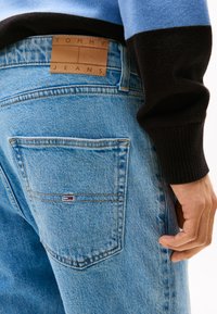 Lyseblå denimjeans med en brun lærlabel som leser "TOMMY JEANS" på midjebåndet og en liten logo på baklommen.