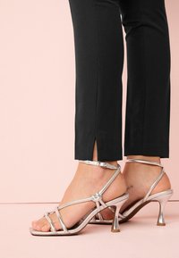Silberne Riemchen-Absatzsandalen mit quadratischer Zehenpartie, schlankem Knöchelriemen und metallischem Finish, kombiniert mit schwarzen Hosen mit seitlichen Schlitzen.