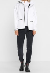 Veste blanche imperméable avec des zips noirs, deux poches avant et une capuche réglable, associée à un pantalon gris foncé et des bottes en caoutchouc noires.