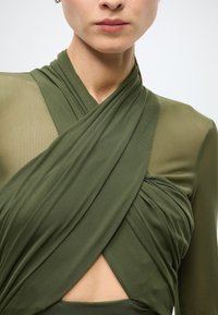 Vêtement vert olive à manches longues avec tissu drapé et accents transparents. Présente un design enveloppant et un détail découpé près de la taille.