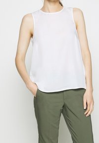 Mujer vistiendo una blusa sin mangas blanca y pantalones verde oliva, con una mano en el bolsillo y un fondo neutro.