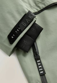 Lichtgroene jas met een gladde textuur, zwart klittenbandpatch met het "BAKER" logo en zwarte ritssluitingdetail op de mouw.