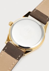 Montre dorée avec un boîtier rond, un bracelet en cuir marron et un dos métallique lisse. Gravure notable sur la partie inférieure.