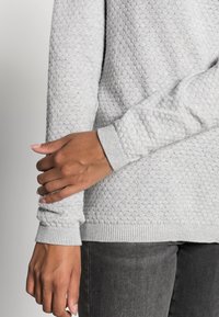 Persona che indossa un maglione grigio chiaro con texture e jeans grigi scuri, con una mano che tiene delicatamente il gomito opposto contro uno sfondo neutro.