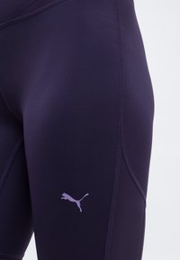 Κοντινό πλάνο ενός ατόμου που φοράει στενά αθλητικά κολάν σε σκούρο μωβ χρώμα με λογότυπο Puma σε ανοιχτό μωβ στην μηρό.