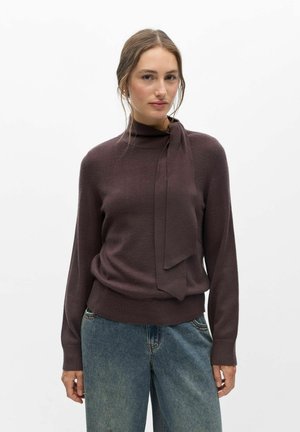 Femme portant un pull marron foncé avec un détail noué au cou et un jean bleu ample, debout devant un fond blanc uni.