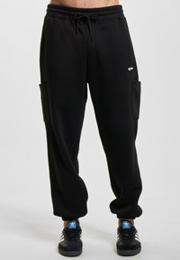 Sorte joggebukser med snor i livet, tettsittende mansjetter, sidelommer og teksturerte detaljer langs yttersemmene.