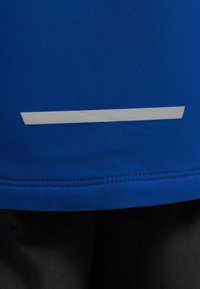 Μπλε αθλητικό τοπ με λεία υφή, που διαθέτει μια αντανακλαστική ασημί οριζόντια ρίγα και ραμμένες λεπτομέρειες στα τελειώματα.