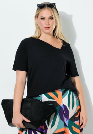 Blondynka w czarnej asymetrycznej bluzce, kolorowej spódnicy z geometrycznym wzorem, trzymająca dużą czarną kopertówkę, z okularami przeciwsłonecznymi na głowie.