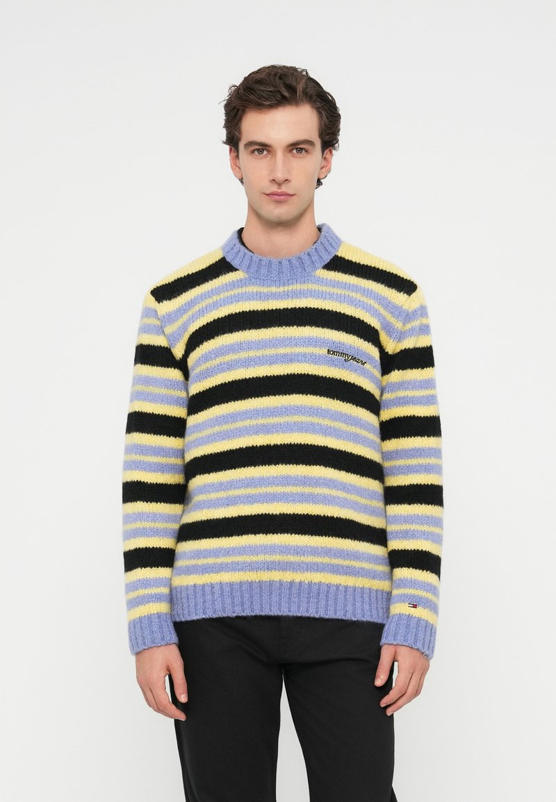 Un jeune homme portant un pull rayé en jaune pastel, noir et lavande, se tenant devant un fond blanc uni.