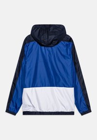 Kapmantel met een blauw en marineblauw ontwerp en een witte band aan de onderkant. Gemaakt van lichtgewicht, gladde stof. Elastische manchetten.