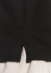 Černé tričko s designem s rozparkem na lemu, vyrobené z měkké látky. Okraje zdobí viditelné švy. Pozadí tvoří světle zbarvená látka.