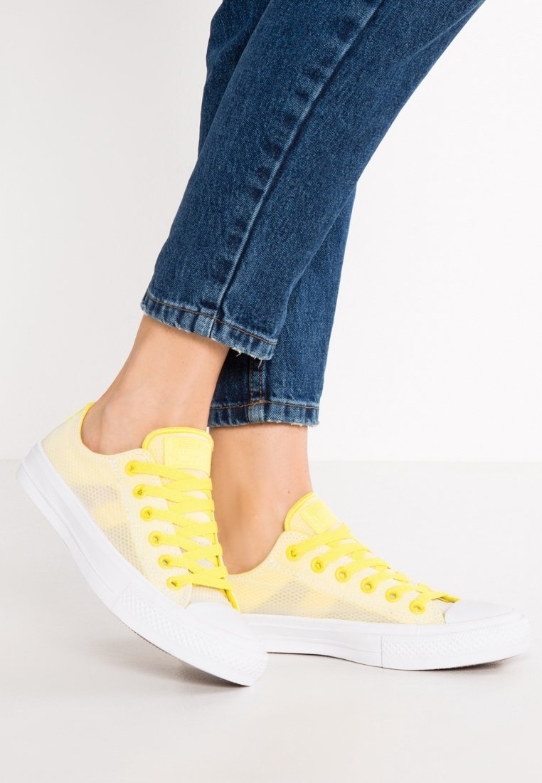 Baskets en mesh jaunes avec des semelles en caoutchouc blanches, dotées de lacets ronds et d'un design basse-toupie, partiellement visibles avec un pantalon en denim bleu.