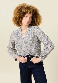 Blusa floreale con maniche lunghe, scollo arricciato, decorata con fiori blu e marroni su uno sfondo chiaro. Abbinata a jeans scuri.
