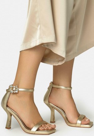 Pies con sandalias de tacón alto doradas con correas alrededor del tobillo y hebillas con piedras brillantes, combinadas con una falda beige.