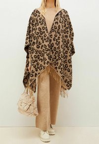 Poncho à imprimé léopard en tissu brun et noir avec un ourlet à franges. Associé à un haut en tricot beige, un pantalon assorti et un sac à main beige duveteux.