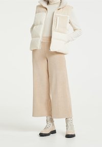Béžový péřový vestový kabát se soft, strukturovaným límcem, vrstvený přes třásňový krémový top a široké béžové kordové kalhoty, doplněný botami světlé barvy.