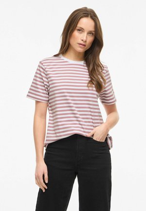 Femme aux longs cheveux bruns portant une chemise à manches courtes rayée rose et blanche et un pantalon noir, debout avec une main dans la poche.