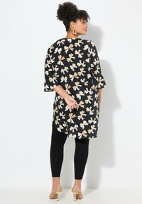Tunic nera a fiori con motivi di foglie beige, abbinata a leggings neri e tacchi nudi. Presenta maniche ampie e una vestibilità rilassata.