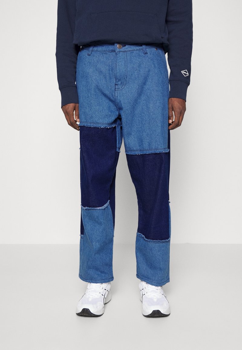 Personne portant un jean patchwork en denim avec des panneaux bleu clair et bleu foncé, un haut à manches longues foncé et des baskets blanches, debout devant un fond uni.